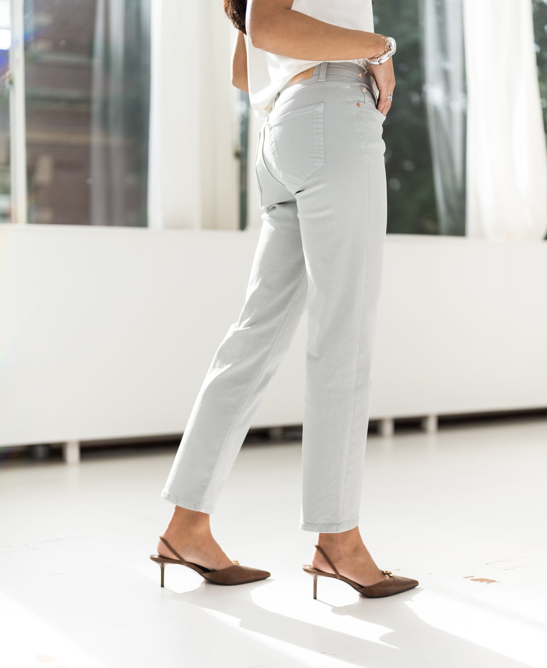 Woman indoors wearing light grey cropped jeans, sunlight streaming in.