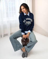 Woman wears PLEIN PUBLIQUE’s LA HARPER dark blue St. Honoré sweater, seated in a bright, minimal room.