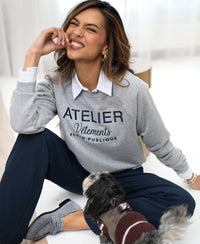Woman wearing a light gray PLEIN PUBLIQUE LA HARPER sweater sits on the floor, smiling.