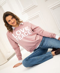 Woman wearing an antique rose knit sweater with LOVE PEACE text sits on floor by a white wall.