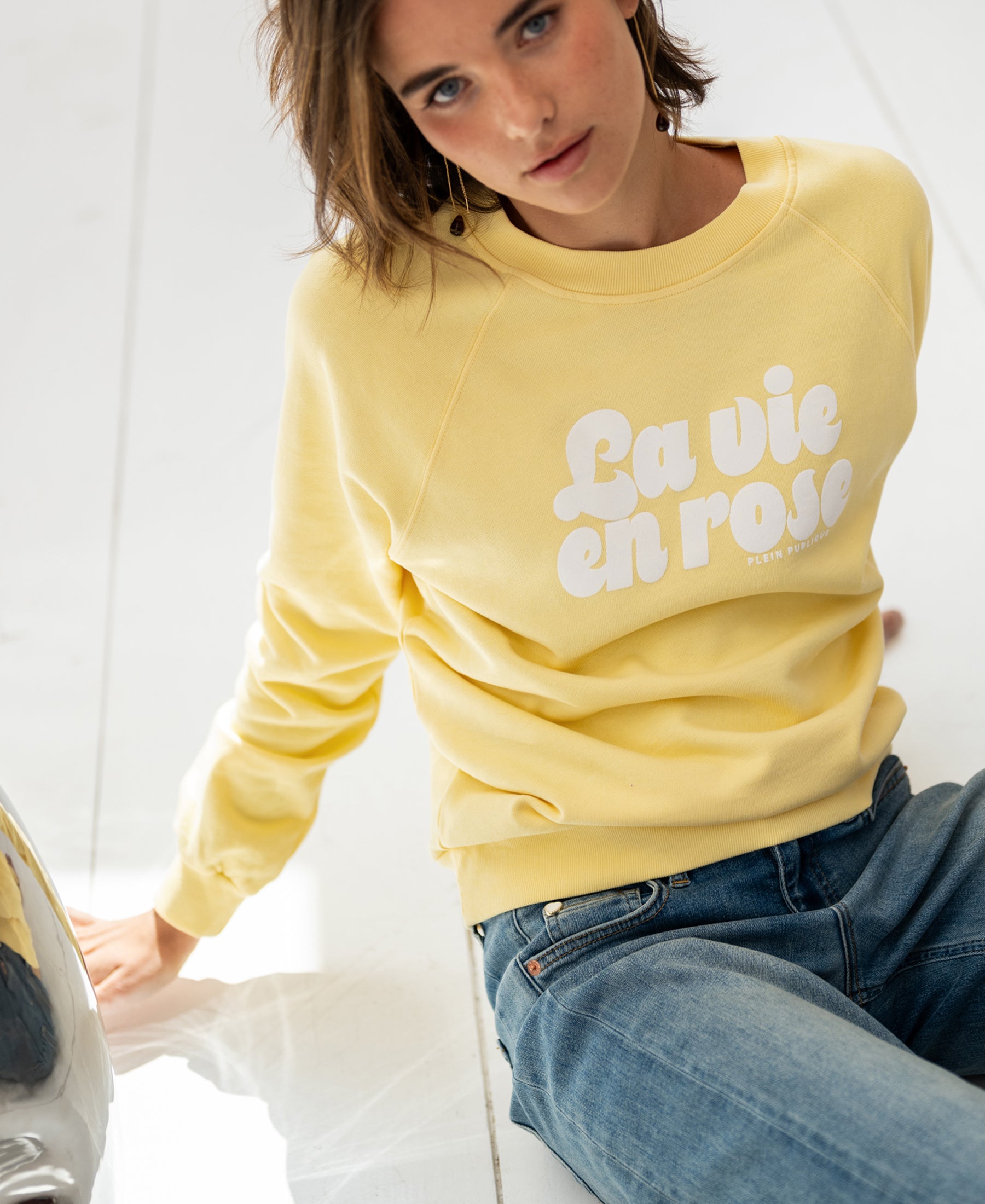 Woman wearing a yellow PLEIN PUBLIQUE LA HARPER sweater sits on a white floor, looking up.