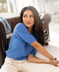 Woman wearing a lavender argyle merino sweater sits by a brown sofa.
