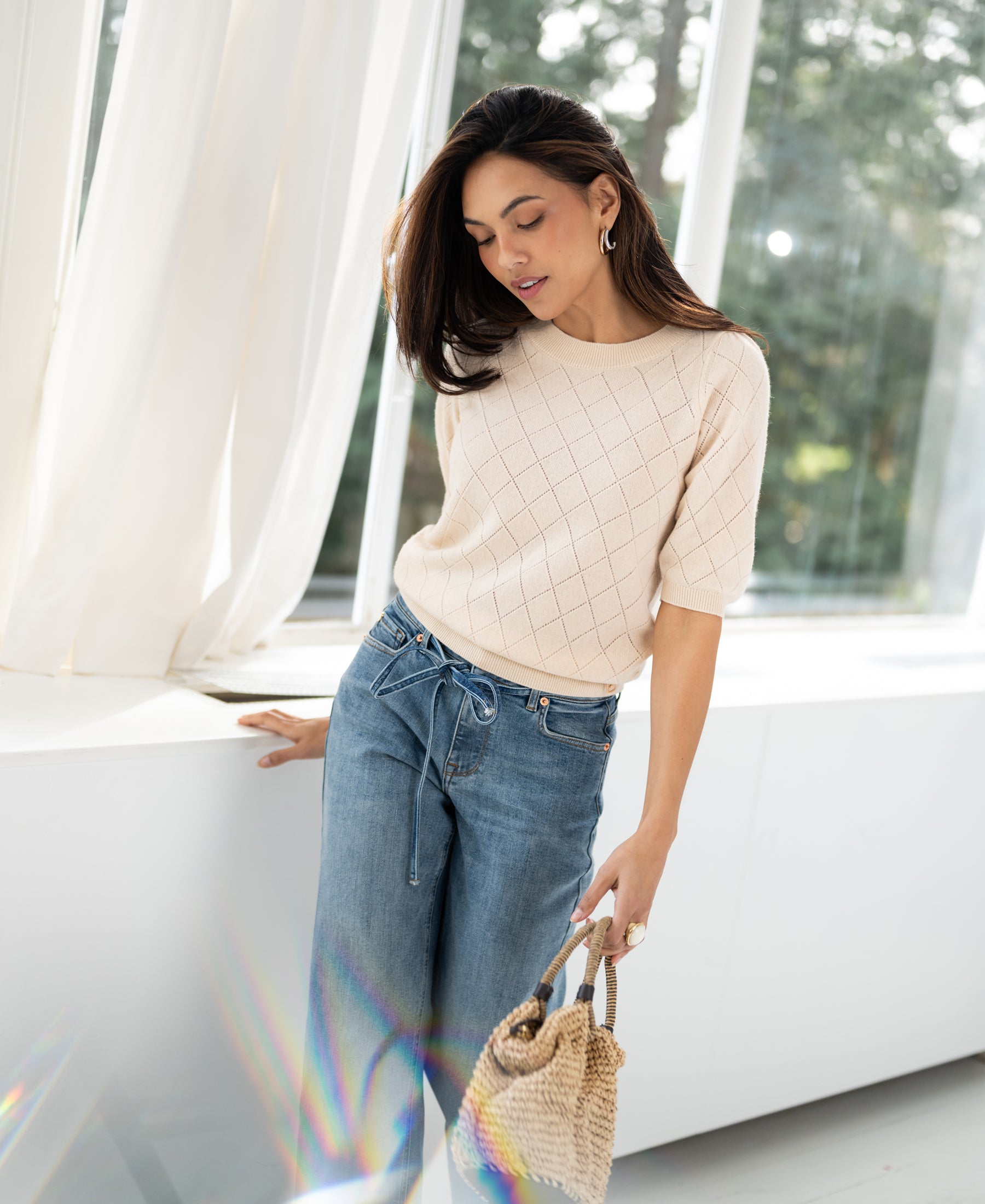 Woman wearing the LE HARVARD Butter argyle merino wool sweater stands by a sunlit window.