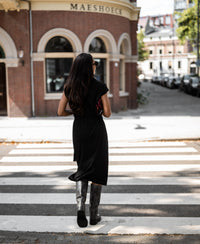 Woman wearing a black PLEIN PUBLIQUE Ines travel polo dress crossing a street with a brick building in the background.