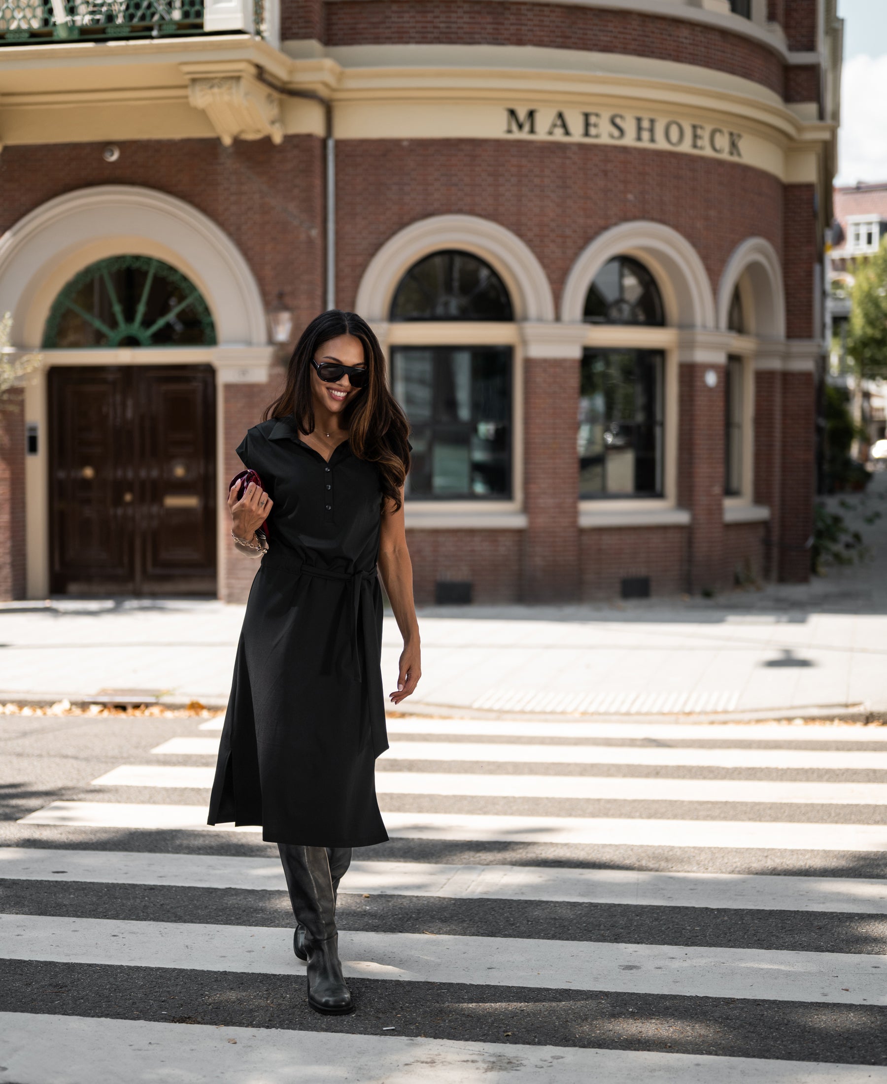 Woman wearing a black INES Travel polo dress by PLEIN PUBLIQUE stands outside a building.
