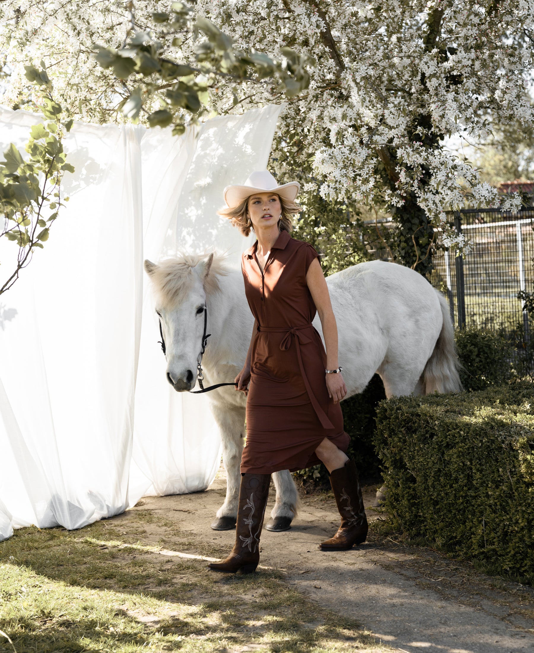 A woman wears a PLEIN PUBLIQUE Travel polo dress INES in camel under blooming trees in bright sunlight.