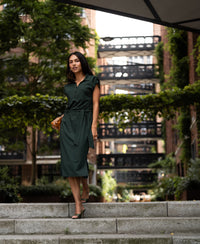Woman wearing a dark green PLEIN PUBLIQUE Travel polo dress walks down steps in a garden with greenery.