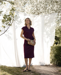 Woman in a wine-colored Travel polo dress stands outdoors with trees and white curtains in the background.