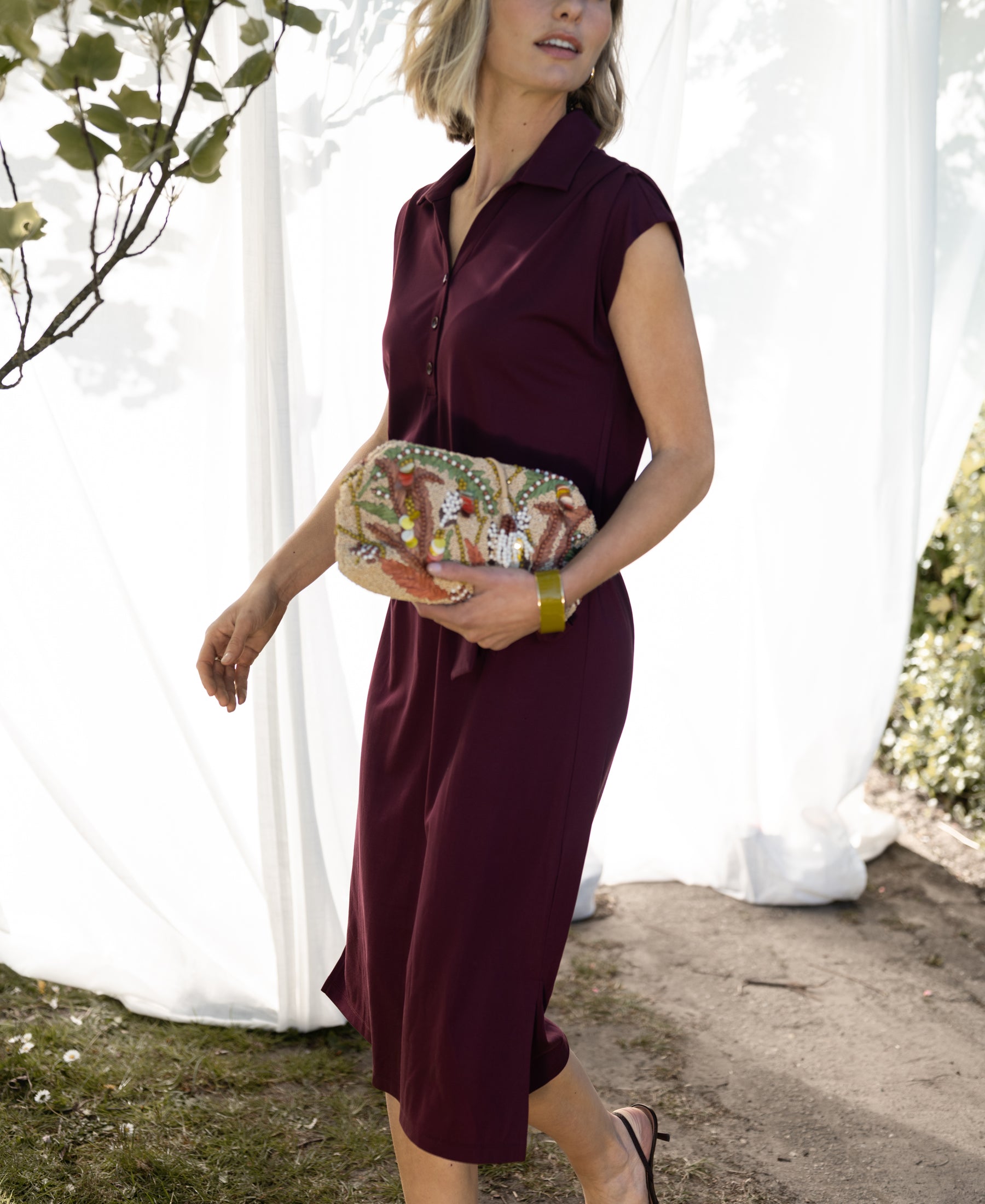 Woman wearing a wine-colored travel polo dress by PLEIN PUBLIQUE walking outdoors by white curtains.