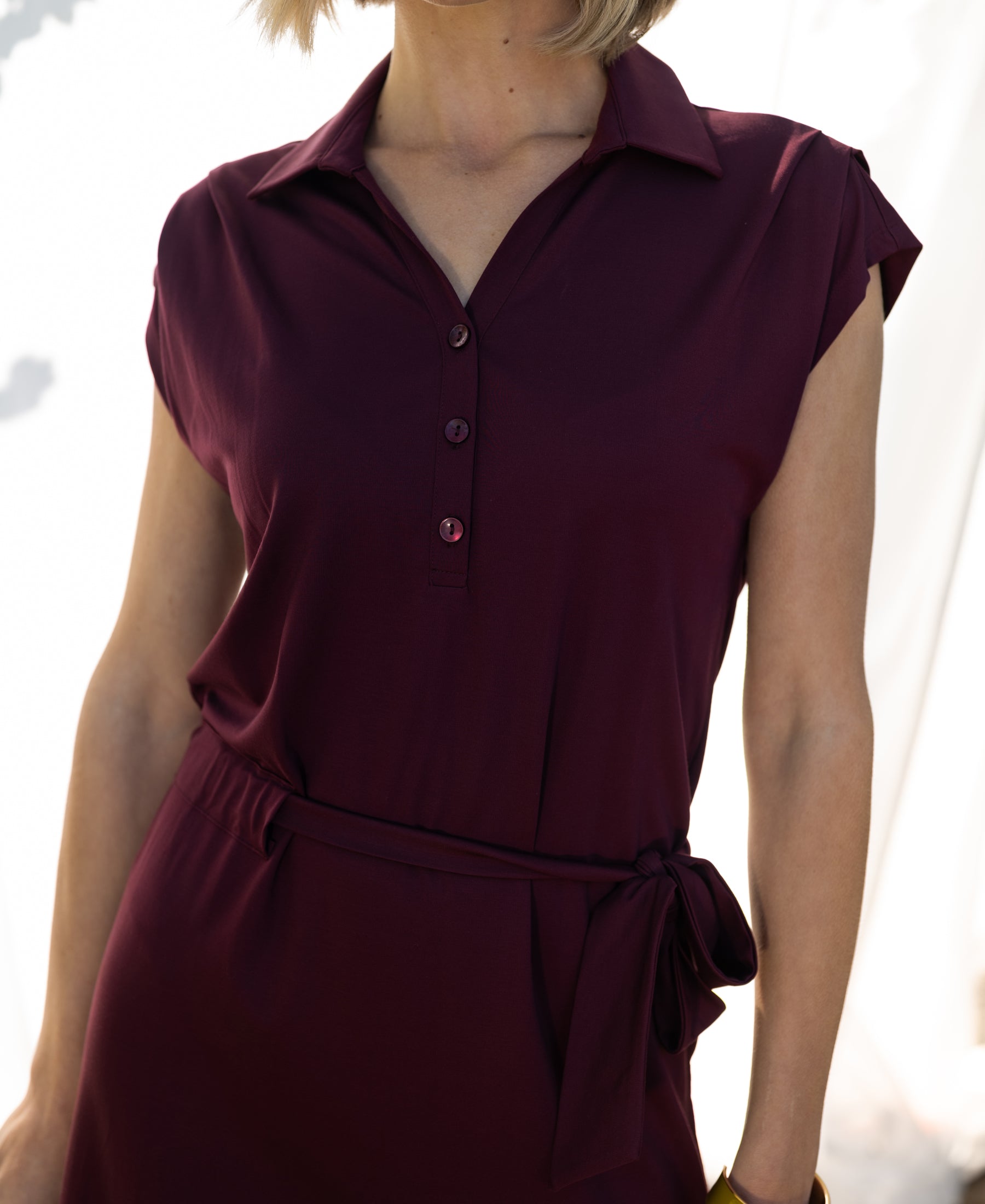 Wine-colored Plein Publique travel polo dress with buttoned front and tied waist, shown in bright light.