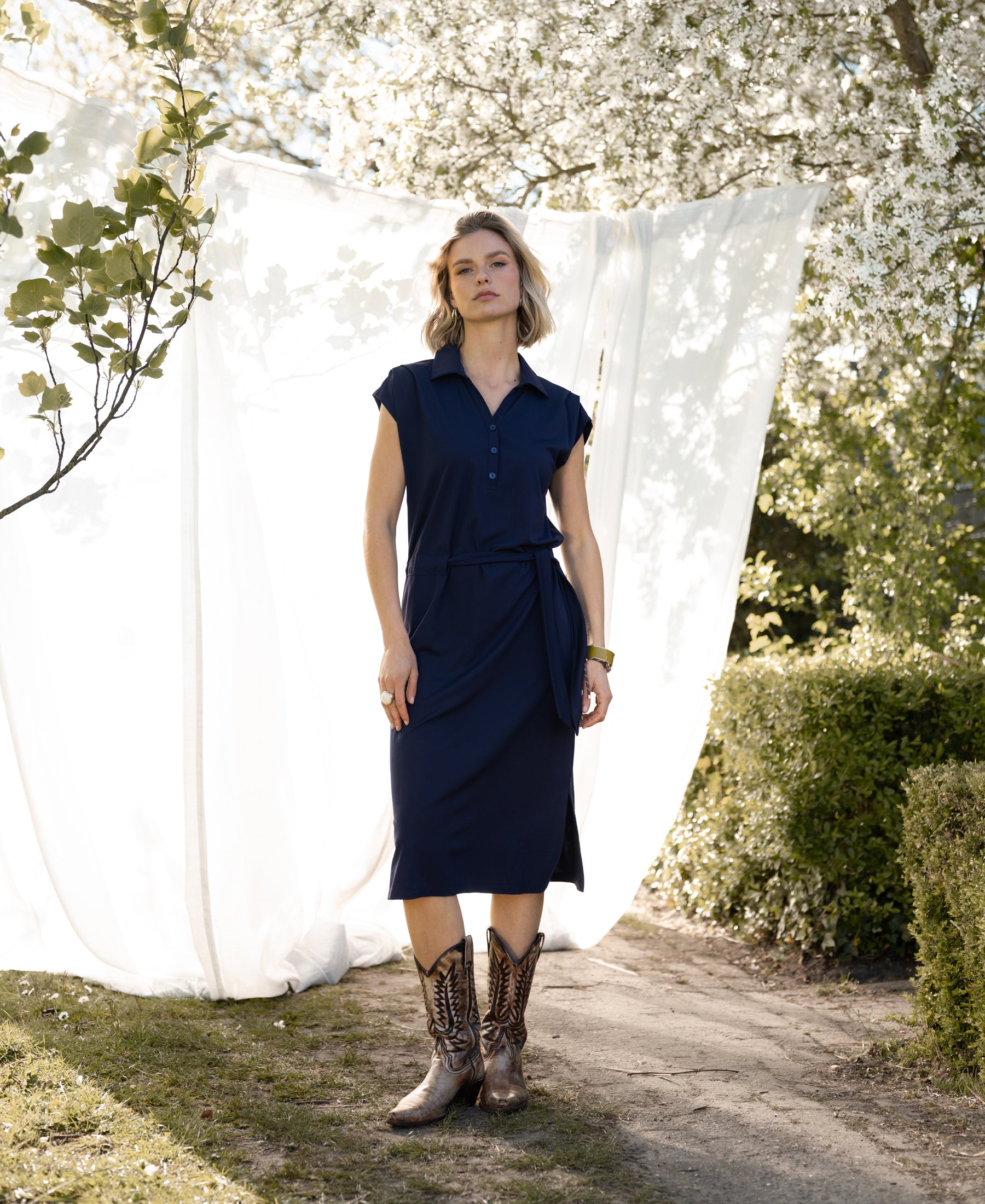 Woman wearing PLEIN PUBLIQUE Travel polo dress INES Dark Blue, standing outside near white curtains and blossoming trees.