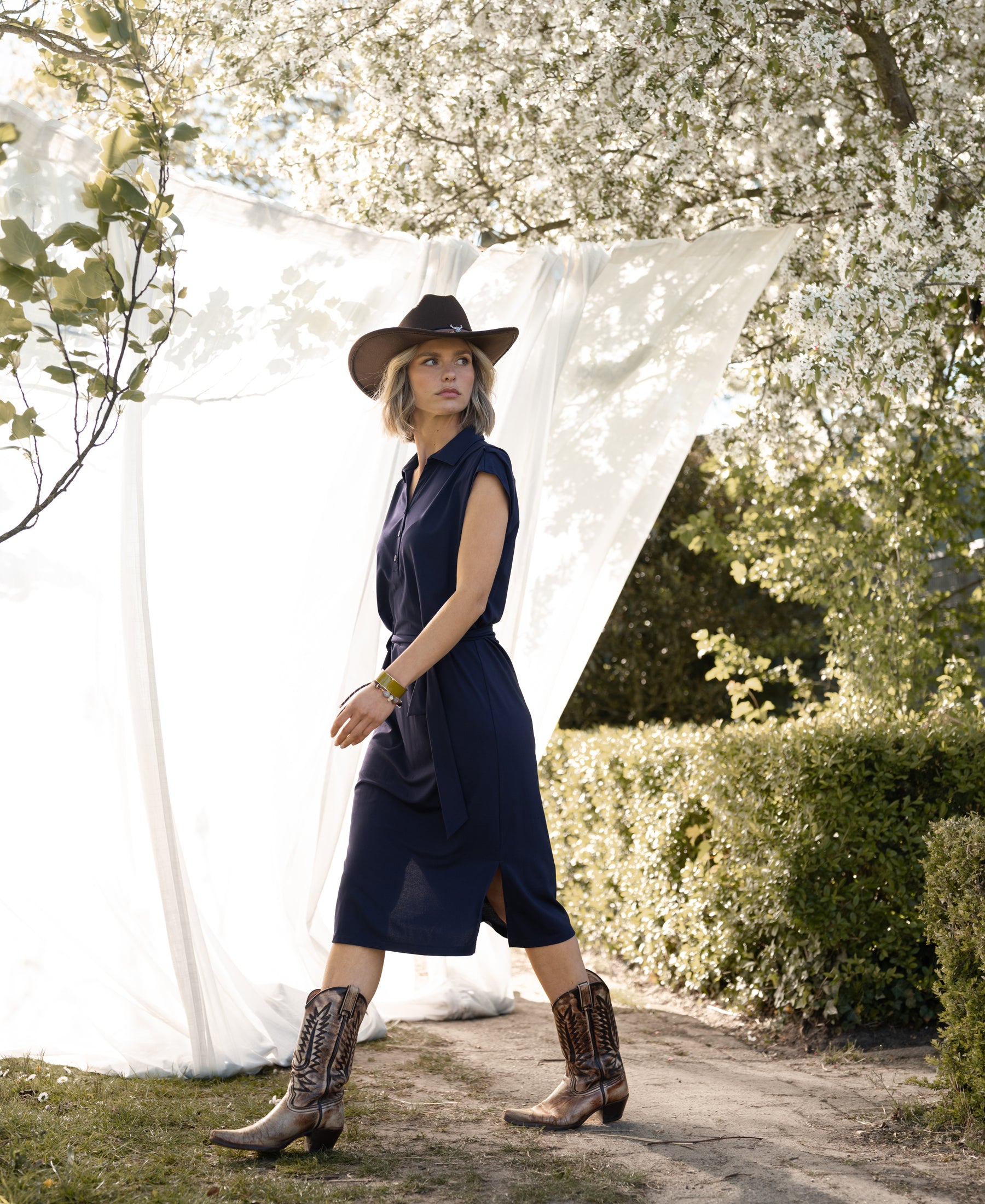 Woman wearing a navy blue PLEIN PUBLIQUE Travel polo dress INES walks outdoors near blooming trees.