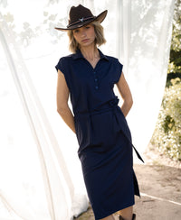 Woman wearing dark blue travel polo dress standing outdoors with white curtains in the background.