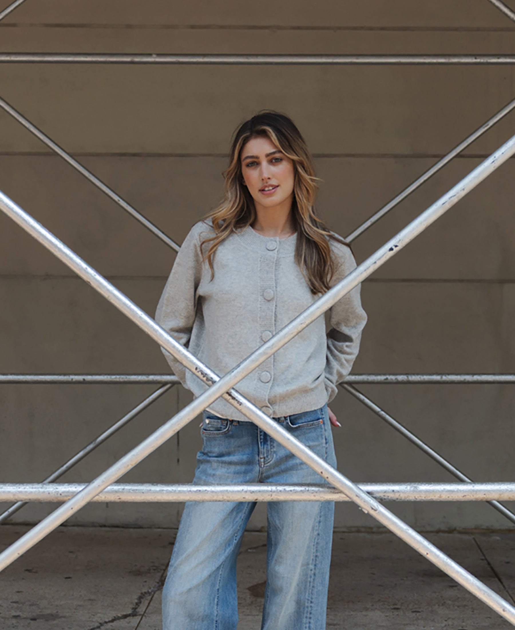 Grey merino wool vest shown on model standing behind crossed scaffolding, plain wall in background.