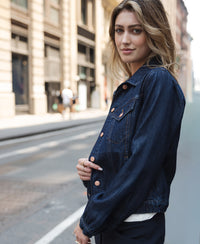 Woman wearing PLEIN PUBLIQUE’s LA JACKY denim jacket in rinse wash, seen standing on a city street.