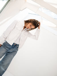 Woman wearing a PLEIN PUBLIQUE LA JACKY white denim jacket, standing indoors, looking down and touching her hair.