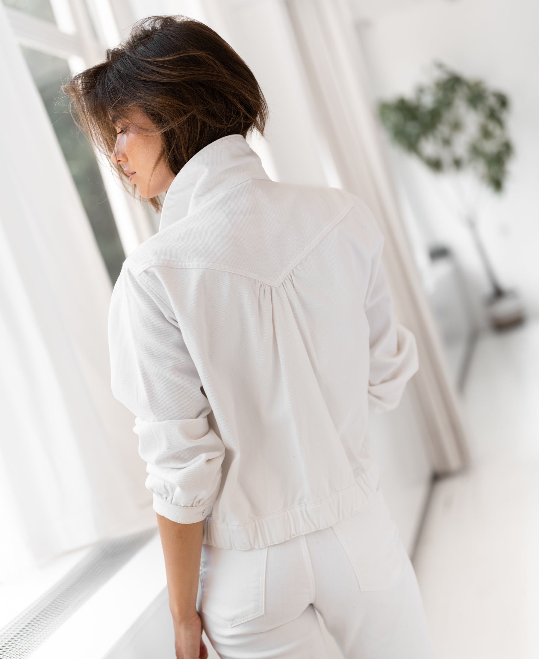 White denim jacket shown indoors by a window with white curtains and a plant in the background.