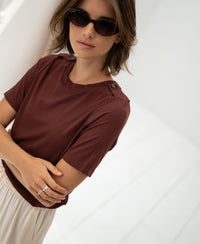 Bamboo T-shirt in red-brown from Plein Publique, arms crossed, shown in a bright white room.