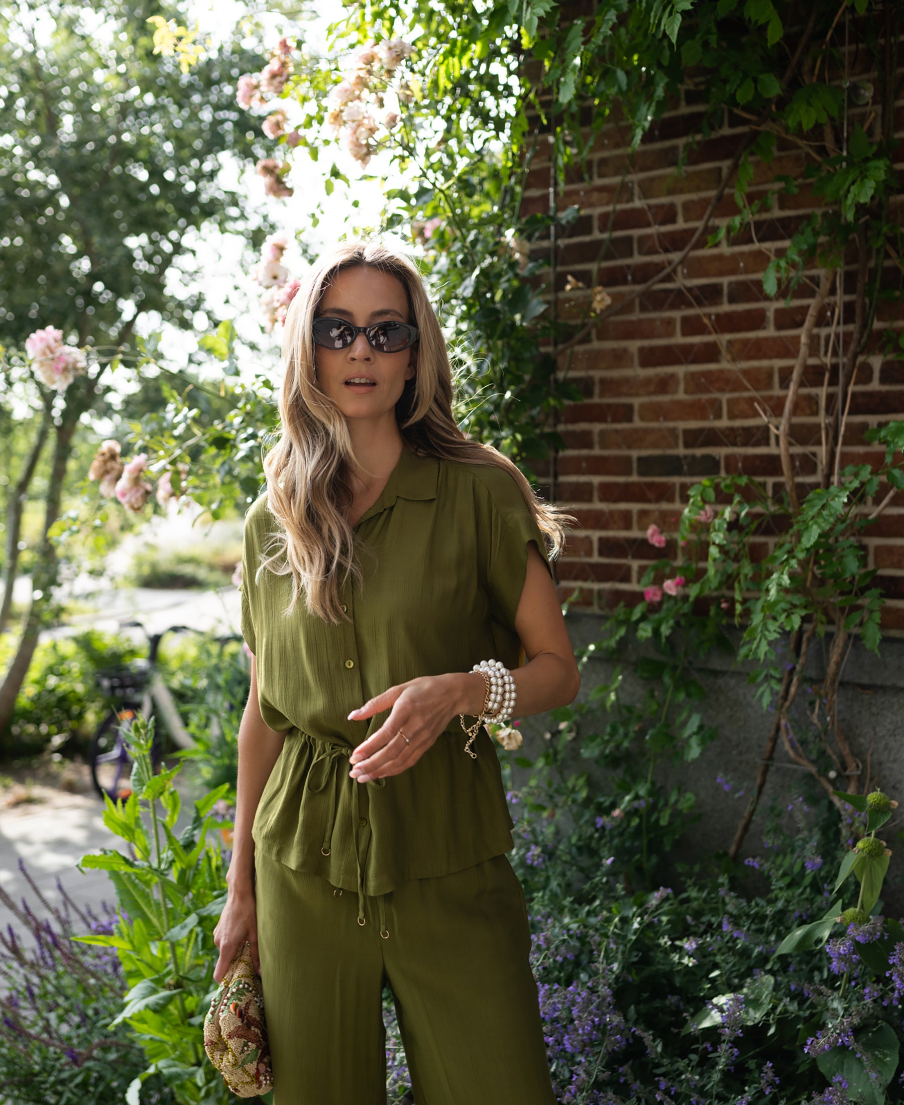 Woman wearing a PLEIN PUBLIQUE olive green blouse, standing by a brick wall with green plants and pink flowers.