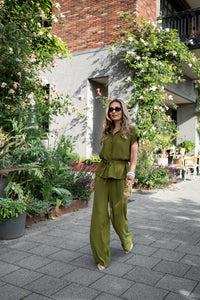 The LA JODY olive green blouse by PLEIN PUBLIQUE displayed outdoors with city greenery in the background.