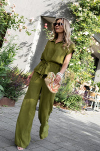 Woman wearing an olive green LA JODY blouse by PLEIN PUBLIQUE, shown outdoors among greenery.