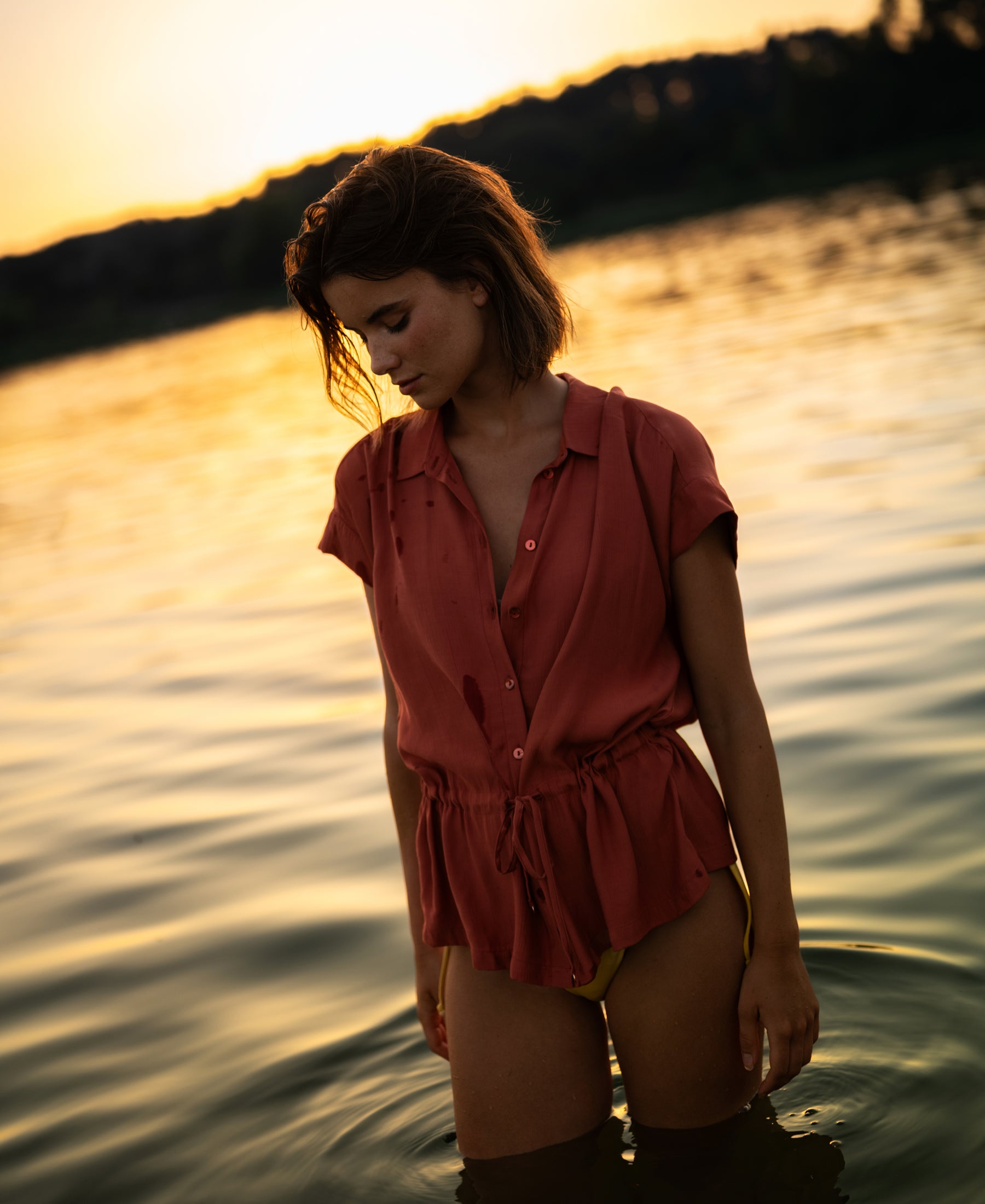 Plein Publique LA Jody terra-colored blouse shown at sunset, displayed on a woman standing in calm water and looking down.