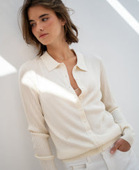 Woman wearing a cream Tencel-silk ribbed cardigan stands against a white wall, looking aside.