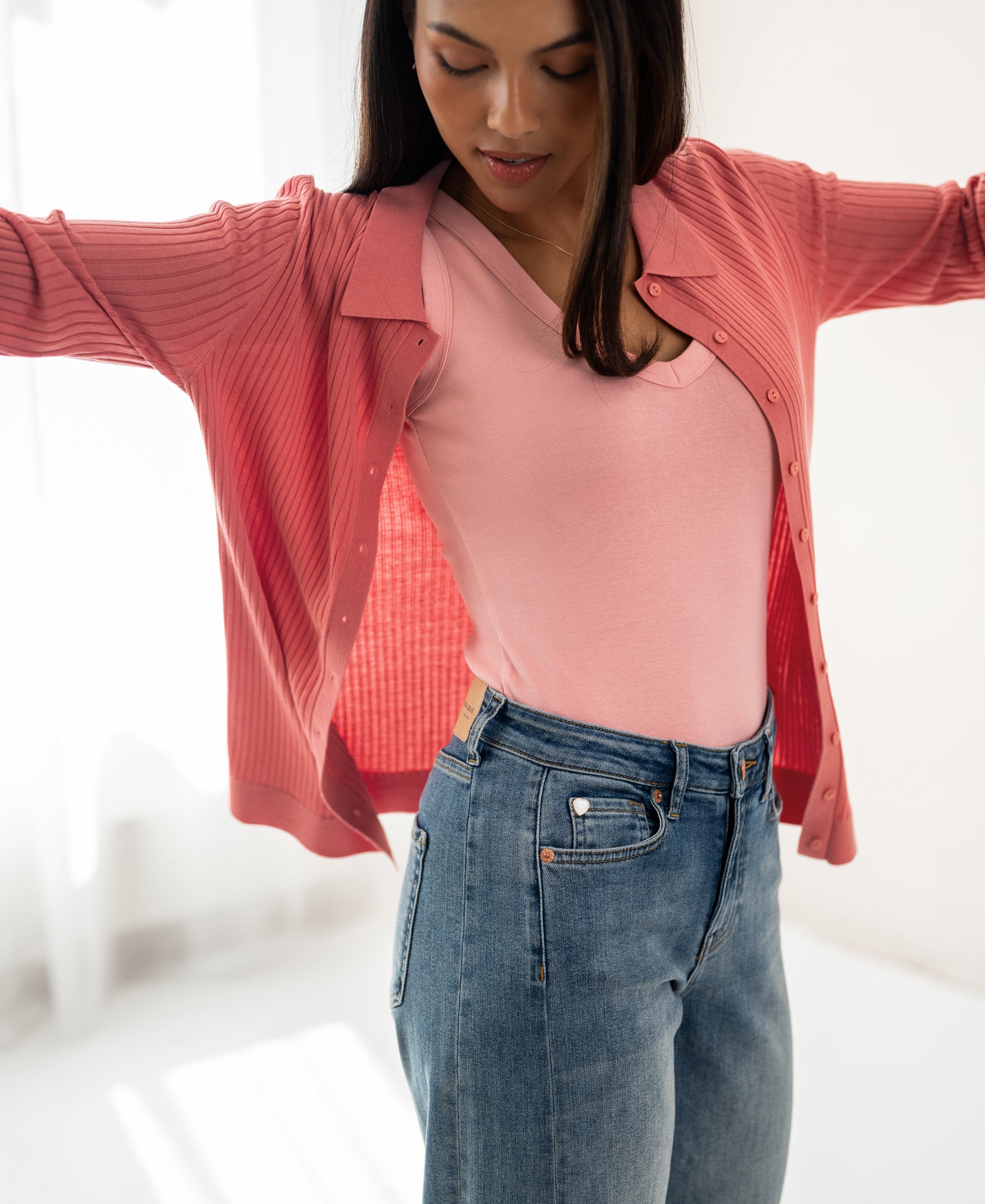 Woman wears pink ribbed cardigan made of Tencel and silk in a sunlit room.