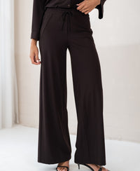 Person wearing the LA JULIA Pure Choco travel pants by PLEIN PUBLIQUE, standing indoors.