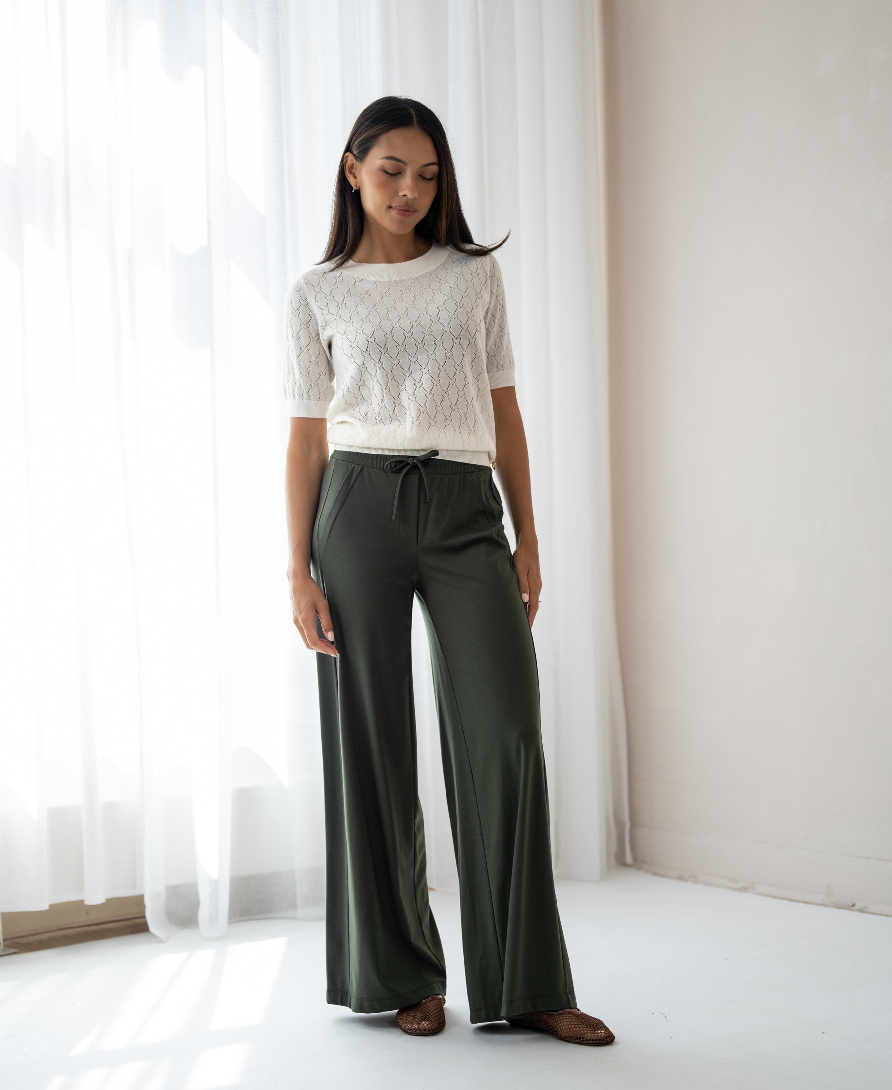 Dark olive Travel pants in a viscose mix, shown indoors by sheer curtains.