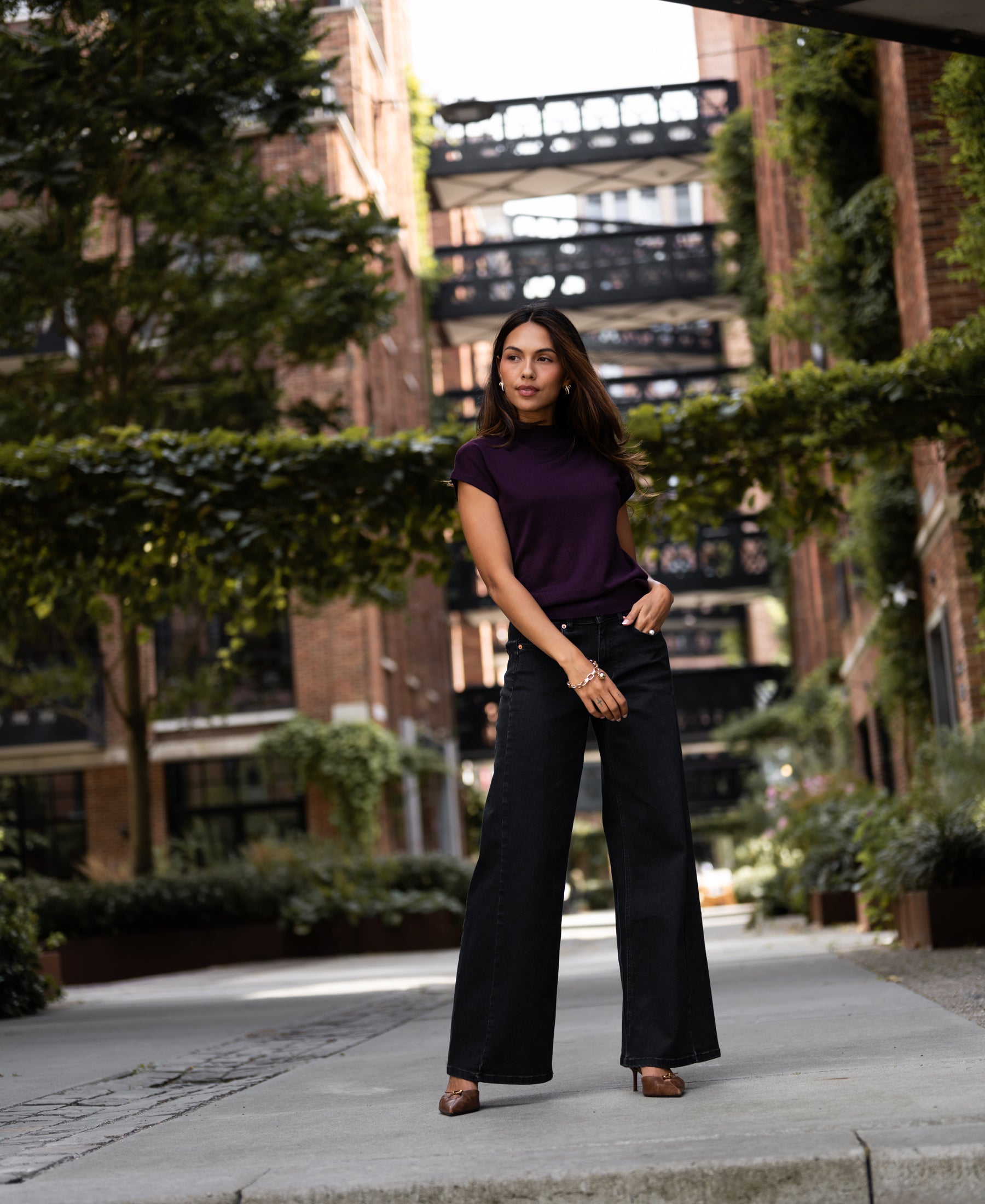Woman wearing a plum Merino wool collared top stands on a city sidewalk with greenery in the background.