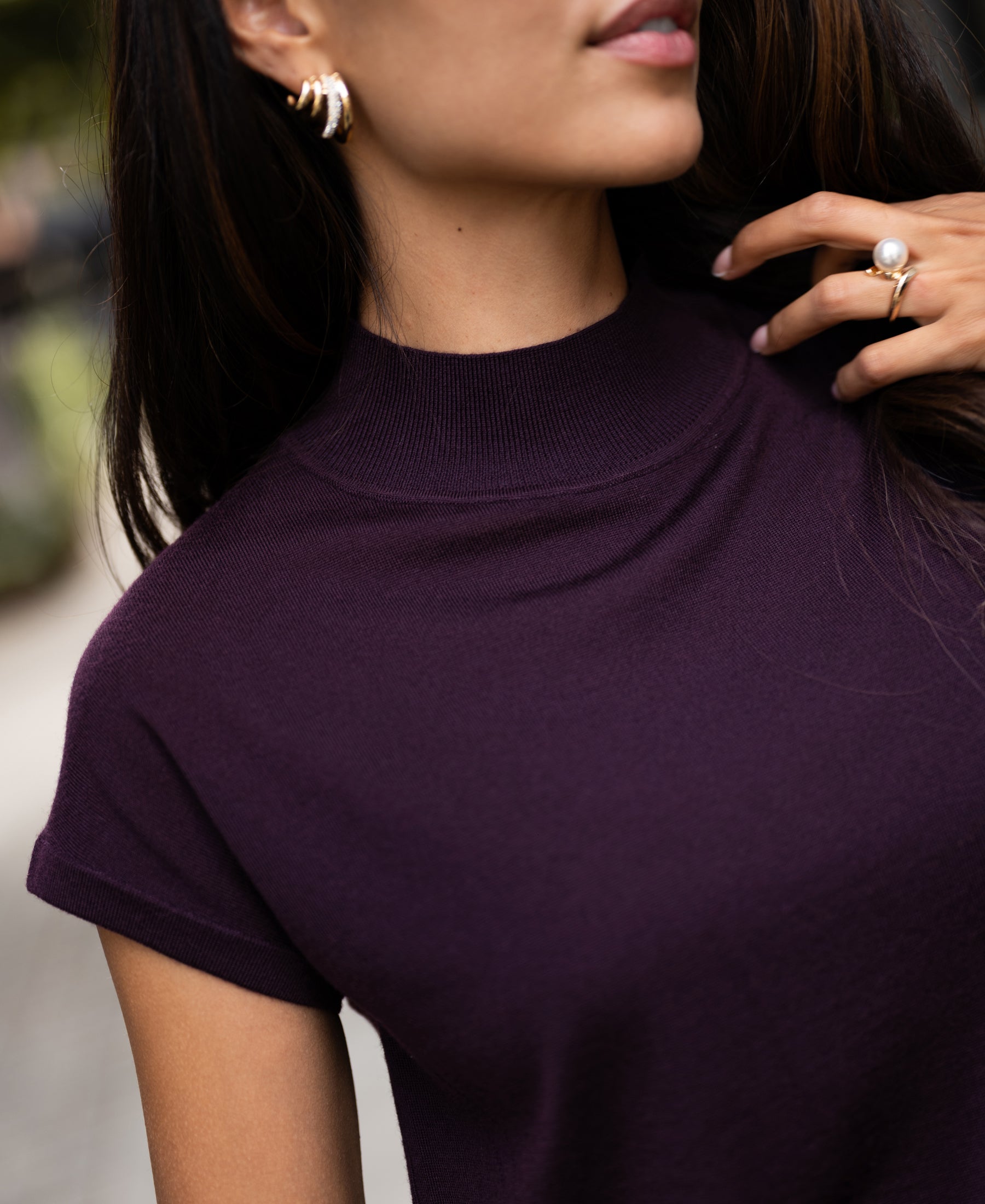 Woman wearing a plum merino wool collared top touches her hair outdoors.