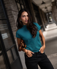 Woman wears the Merino wool LE JULIAN collared top in dark teal, standing outside a brick building.