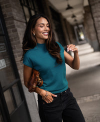 Woman wearing a dark teal merino wool collared top, smiling while standing on the sidewalk.