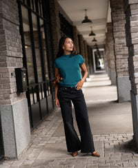 Woman wearing a dark teal Merino wool top stands outdoors, gazing sideways.