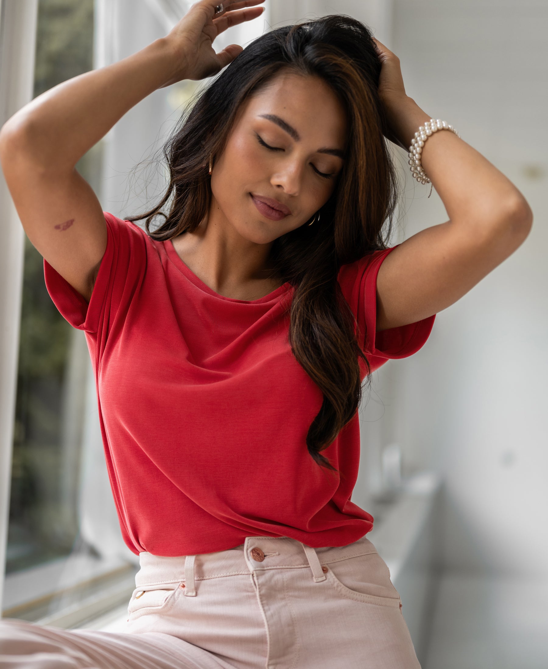 Woman wearing red PLEIN PUBLIQUE two-way T-shirt poses by a window, eyes closed.