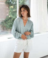Woman wearing a light green and ivory wrap blouse with dot pattern stands by a sunlit window.