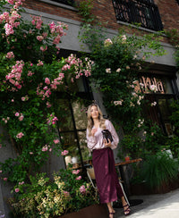 Woman wearing wrap blouse in dusty pink with ivory polka dots stands outside.