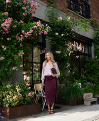 Woman in dusty pink polka dot wrap blouse stands outside a café with pink roses.