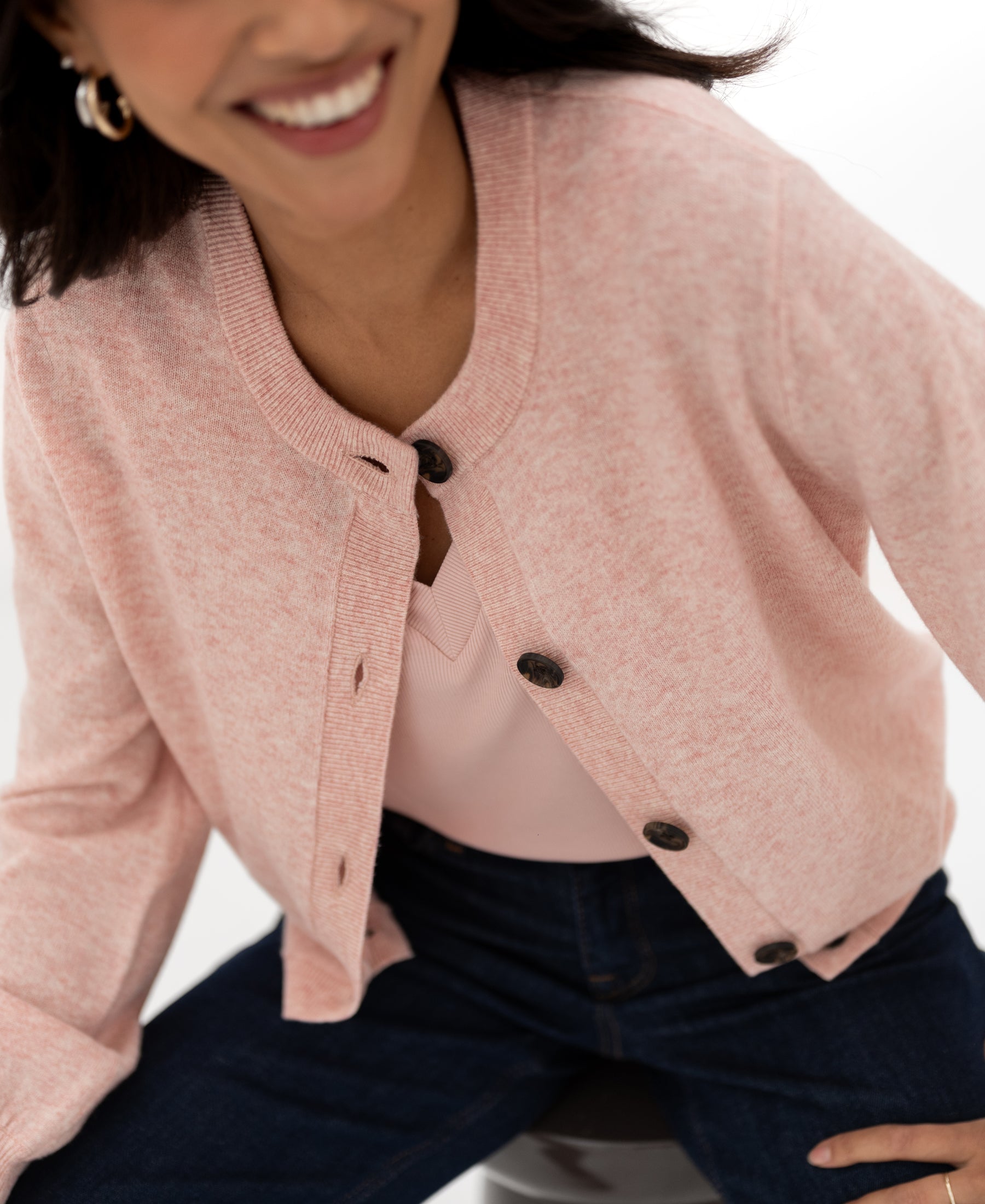 Woman wearing a pink PLEIN PUBLIQUE round-neck cardigan, seated on a stool.