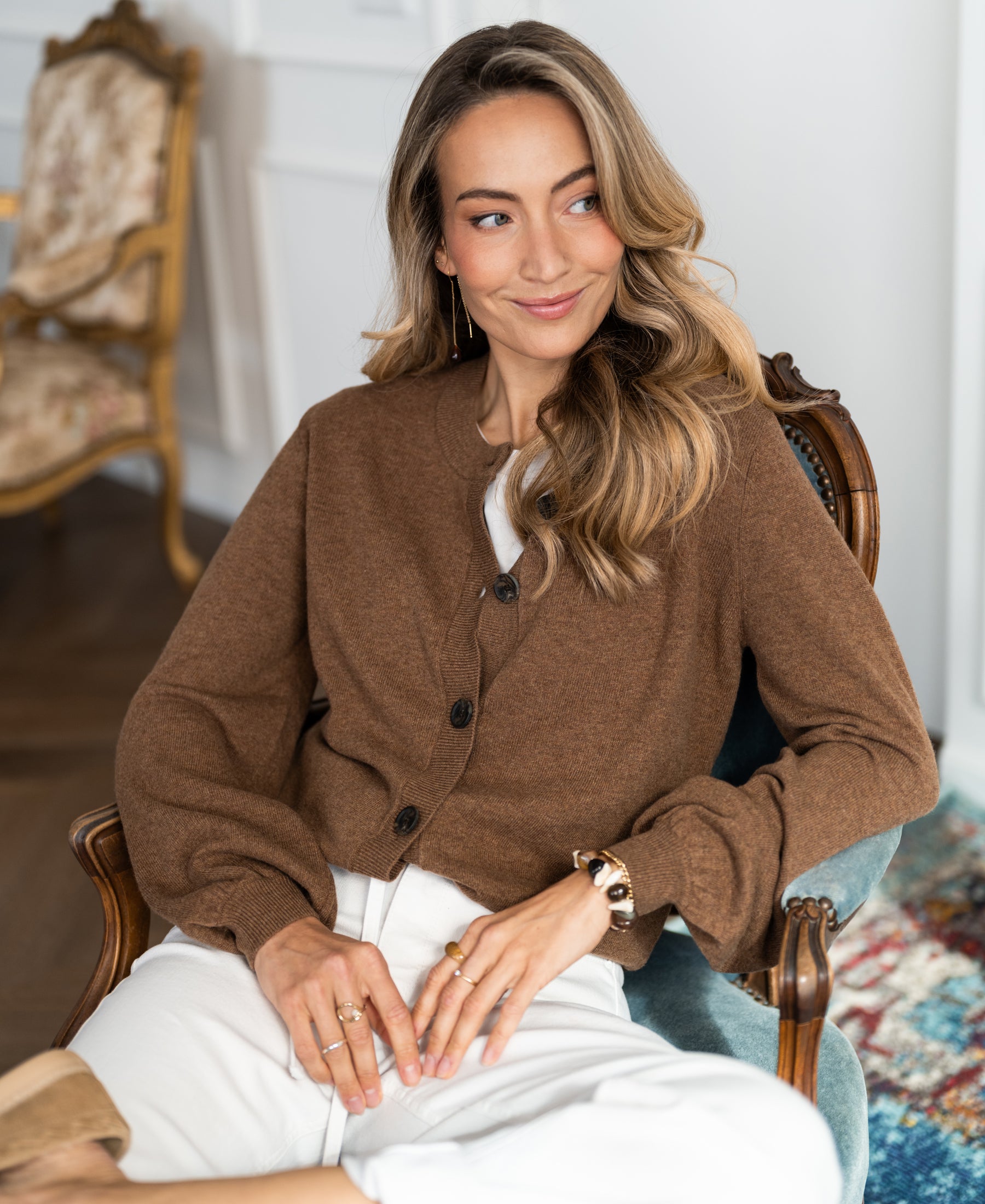 Woman with long, wavy hair in ornate chair wears tobacco-colored round neck vest, smiling.