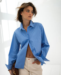 Oversized easy-iron blue blouse shown from the front, with model standing and one hand in her pocket, looking to the side.