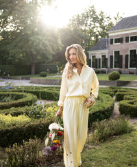 Light yellow LA LYNETTE trousers by PLEIN PUBLIQUE, shown in a sunlit garden with a house in the background.