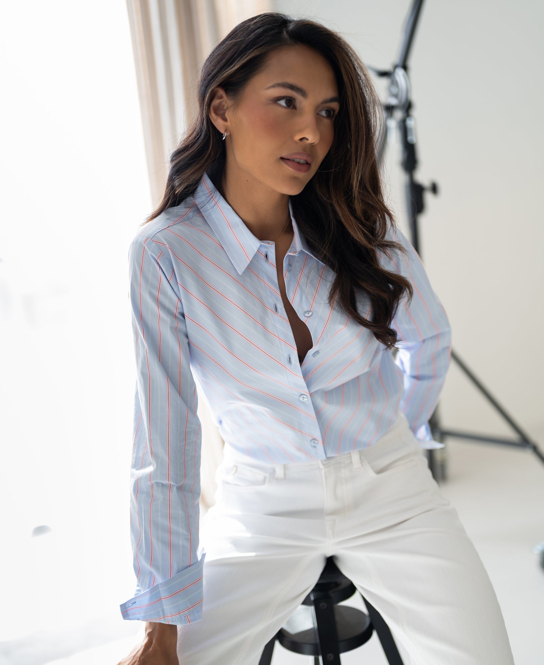 Woman wears striped blouse LA LOIRE in light blue and coral, seated in a modern, bright studio.