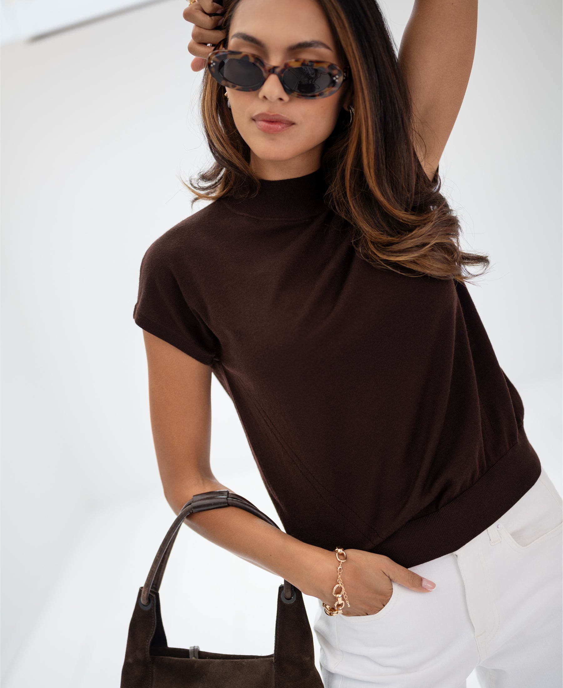 Woman in brown merino wool top holds a matching brown bag against a bright background.