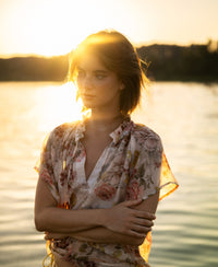 A blush floral print blouse top from PLEIN PUBLIQUE is shown, modeled outdoors near a lake at sunset.
