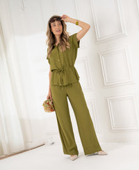 Woman wearing olive green LA LYNETTE trousers by PLEIN PUBLIQUE stands in a bright, white room.