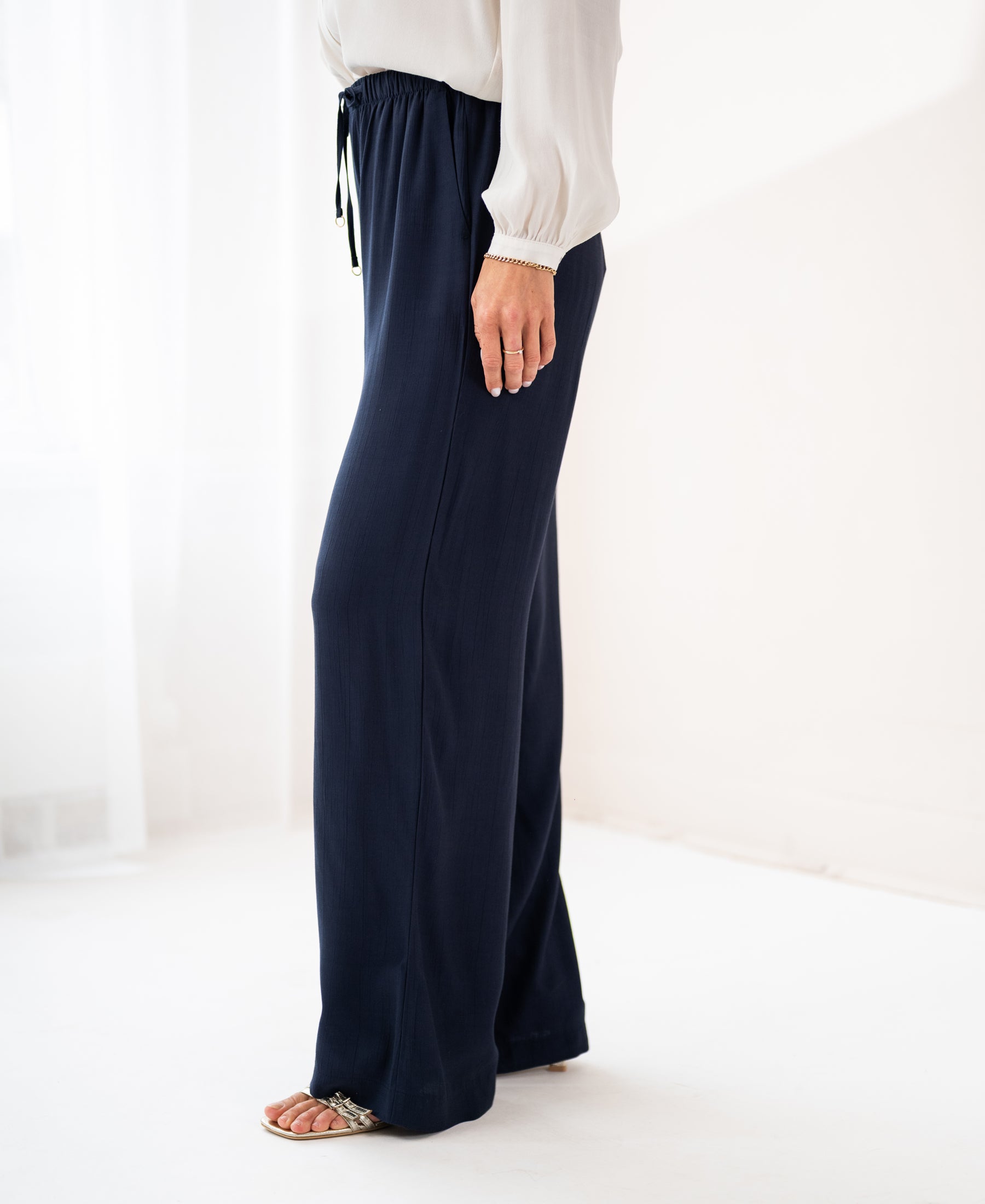 A person wears dark blue LA LYNETTE trousers in a bright, minimal room.