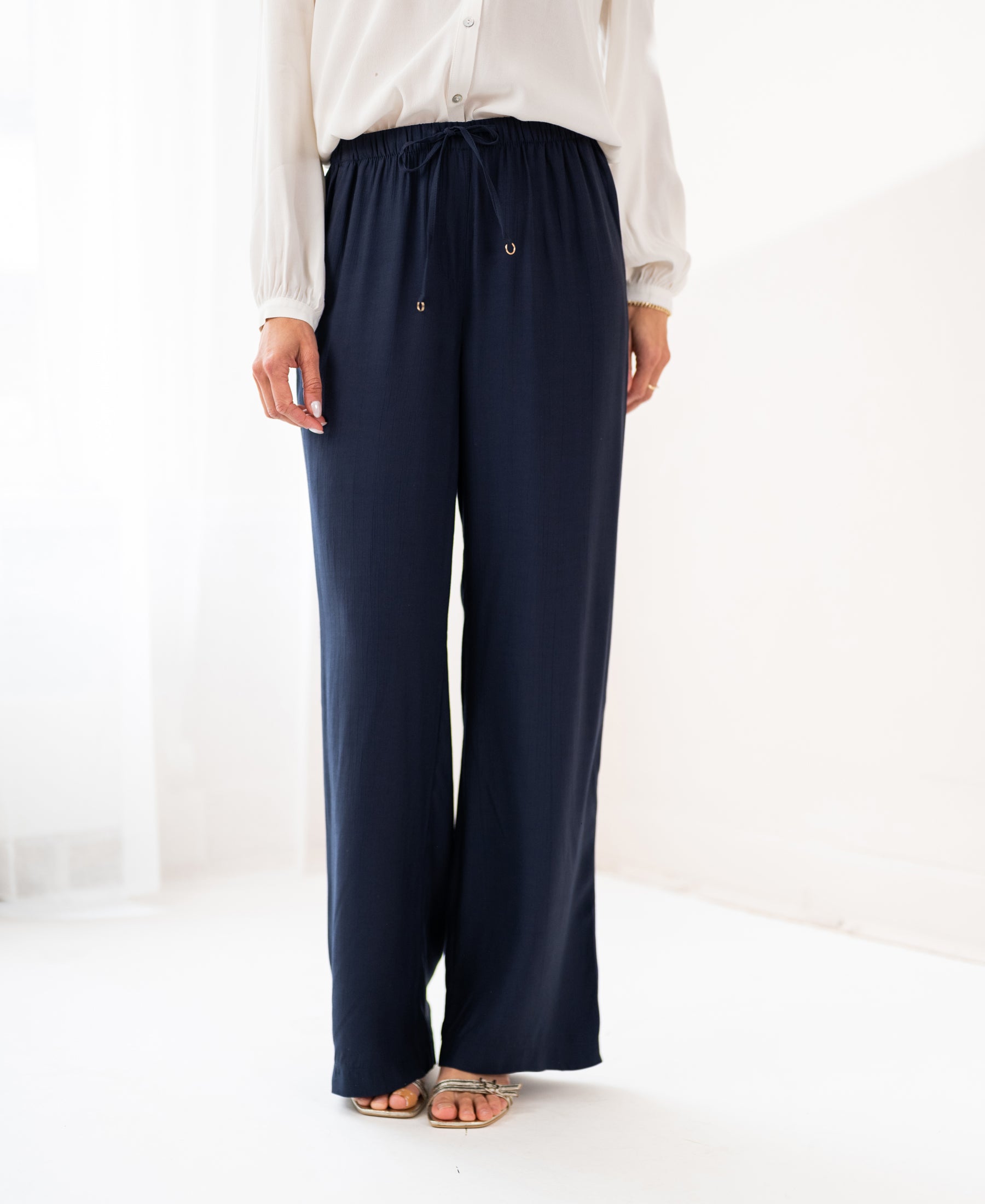 Person wearing dark blue LA LYNETTE trousers by PLEIN PUBLIQUE, standing indoors on a light floor.