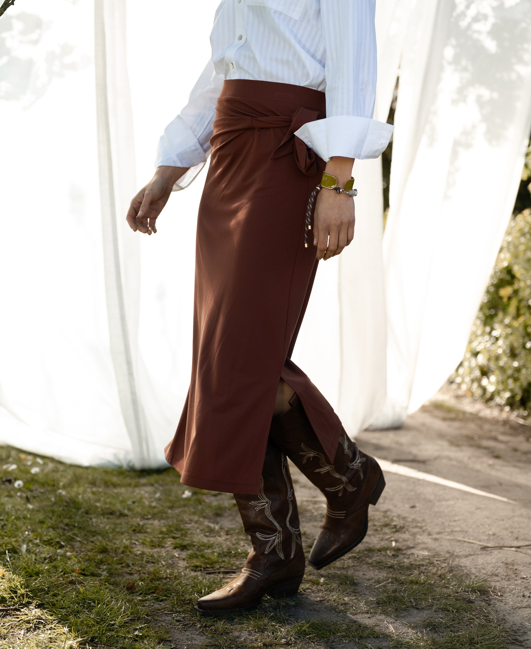 Camel-colored LE MAEL travel skirt by PLEIN PUBLIQUE shown outdoors on grass near white curtains.